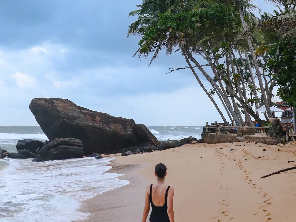 Mariana in a black swimsuit walks along a serene, golden beach in Sri Lanka, with dramatic rock formations and leaning palm trees under a moody sky. This peaceful coastal scene captures the kind of hidden gem you might discover on a well-planned 10 day Sri Lanka itinerary.