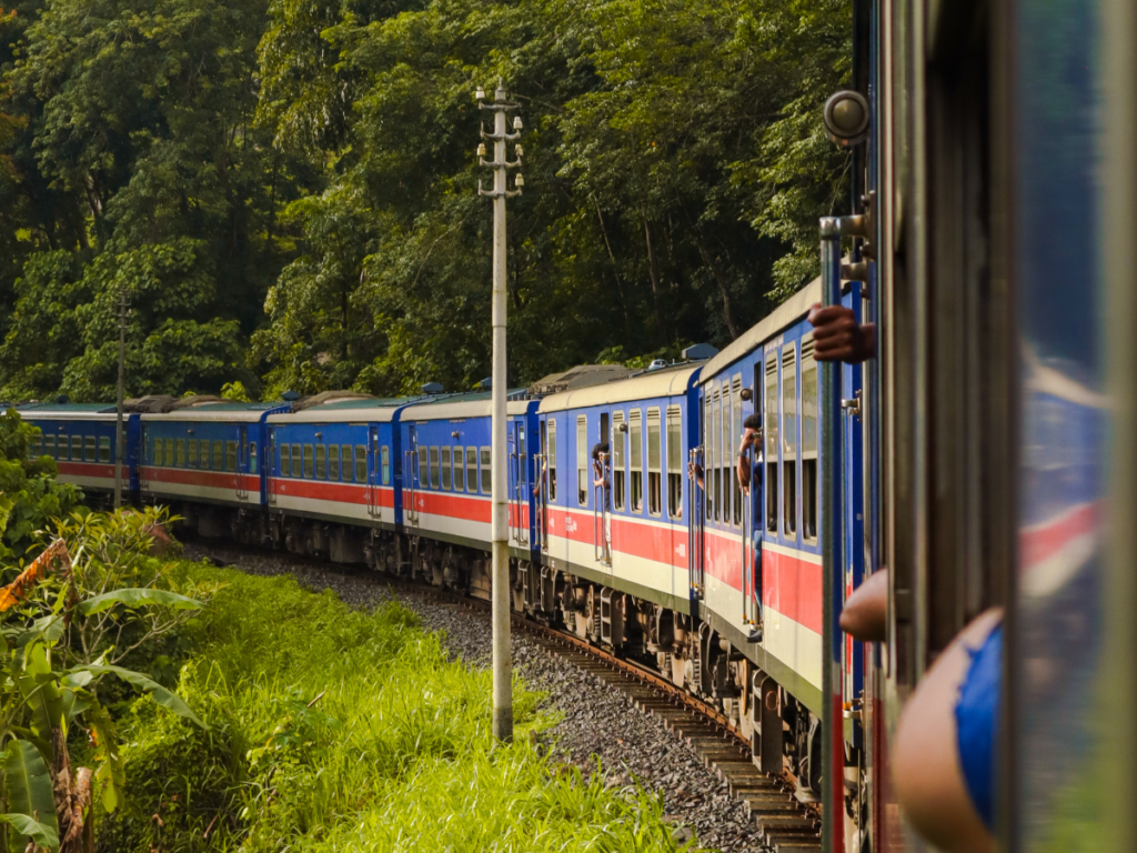 A long blue train with red and white stripes curves through lush green jungle, with passengers leaning out for a better view. This is one of the most scenic stretches of the kandy to ella train ride, loved for its jungle-lined tracks and vibrant carriages.