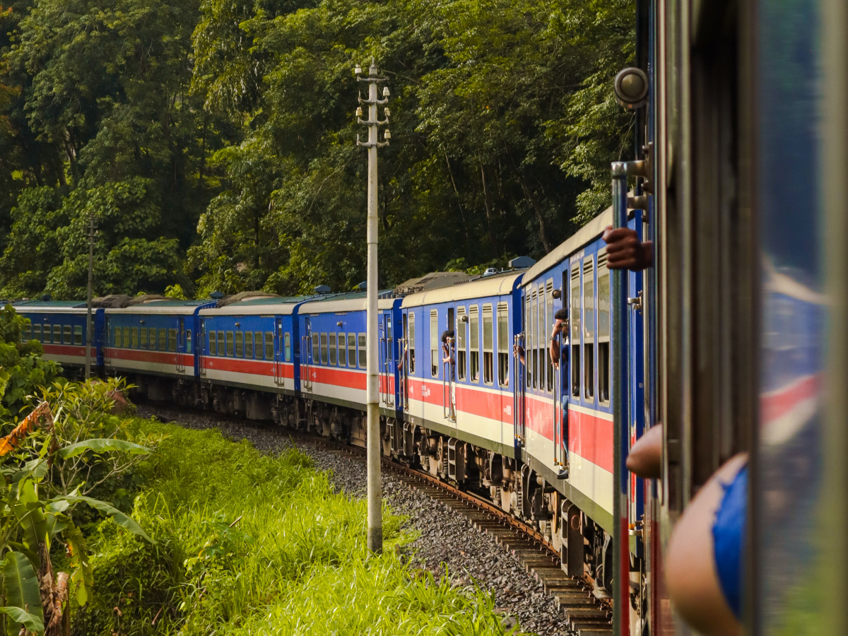 A long blue train with red and white stripes curves through lush green jungle, with passengers leaning out for a better view. This is one of the most scenic stretches of the kandy to ella train ride, loved for its jungle-lined tracks and vibrant carriages.
