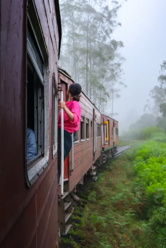 Mariana in a bright pink shirt leans out of a red train as it curves through lush green hills on the iconic Ella to Kandy train ride. The scenic railway hugs the edge of a mountainside, offering a thrilling view of Sri Lanka’s natural beauty.