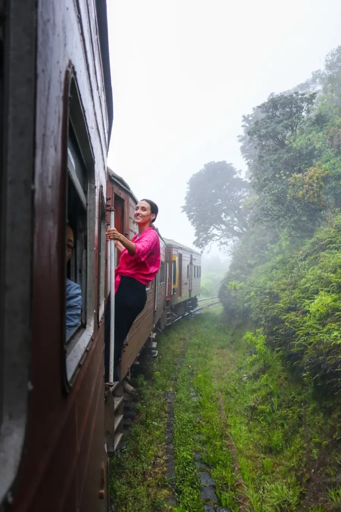 Mariana in a pink shirt leans out from a train door as it winds through misty forest hills. The kandy to ella train offers these immersive open-air moments surrounded by nature.