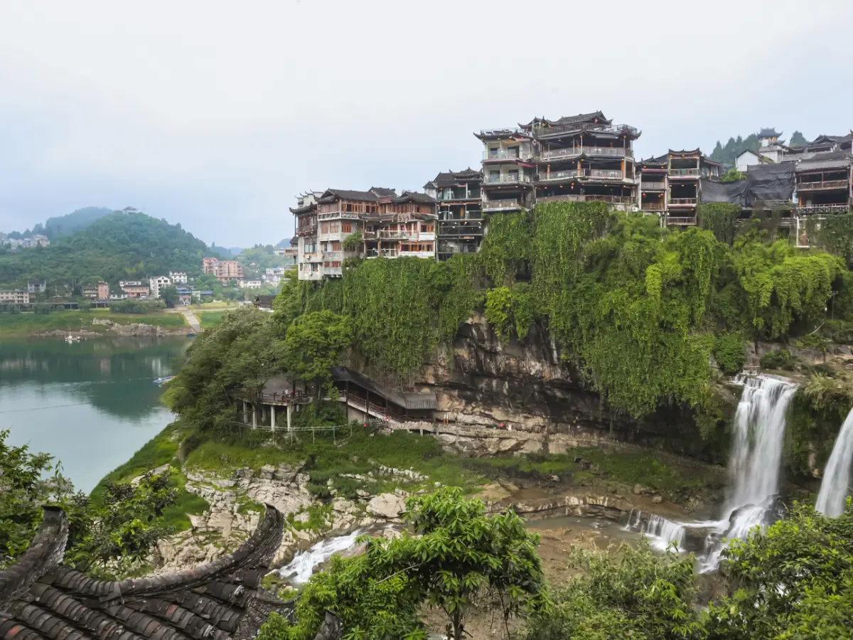 The cascading Furong Waterfall flows beneath historic wooden structures covered in lush greenery; a scenic spot that's iconic to Furong Ancient Town.