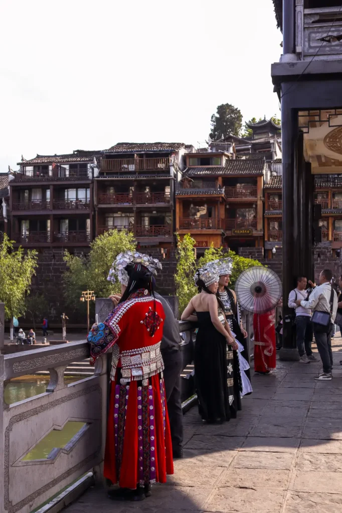 China Travel Tips: 35+ must-know things before visiting (2026 Guide) 13 People dressed in traditional Miao-style outfits with silver accessories stand on a bridge, facing an old town with wooden buildings. A cultural China tip for tourists is to try traditional dress-up experiences in heritage towns.