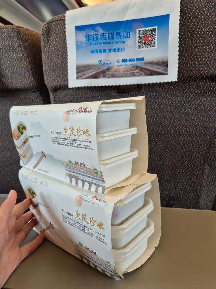 How to buy train tickets in China (2026): easy & tourist-friendly options 10 Two stacked train meal sets in white plastic containers with printed paper wraps are placed on a tray table inside a Chinese high-speed train.