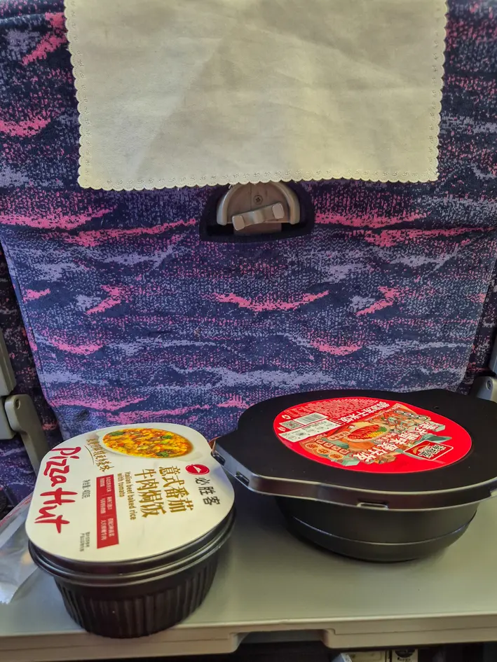 How to buy train tickets in China (2026): easy & tourist-friendly options 9 Two instant meal bowls, including a Pizza Hut beef rice option, rest on a tray table aboard a Chinese train.