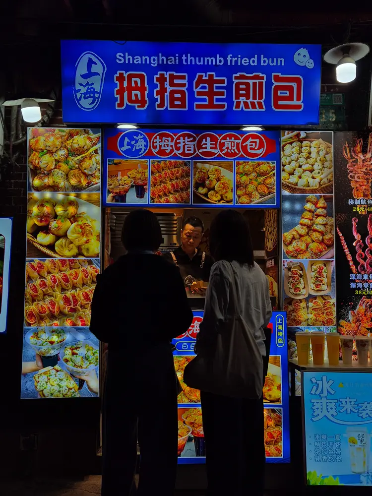 China Travel Tips: 35+ must-know things before visiting (2026 Guide) 18 A bright blue sign reads "Shanghai thumb fried bun" as two women order at a night market food stall with colorful bun photos.