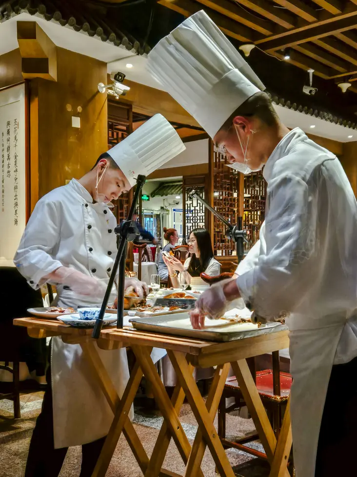 China Travel Tips: 35+ must-know things before visiting (2026 Guide) 16 Two chefs in tall white hats slice Peking duck at a table in front of restaurant diners