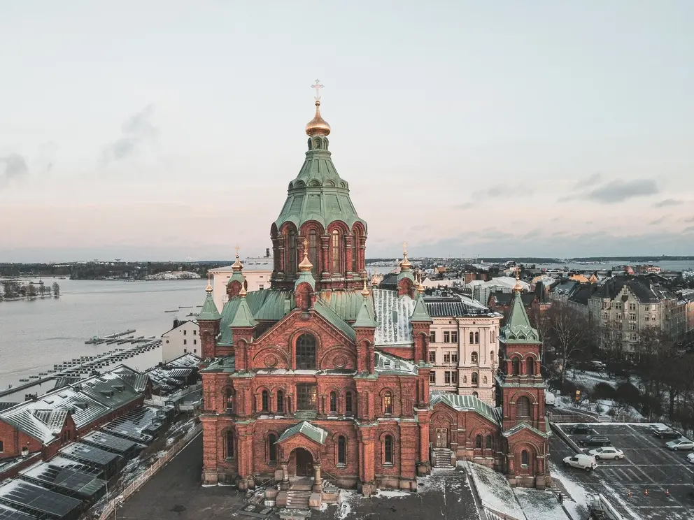 Where to stay in Helsinki: the best neighborhoods & hotels 6 Aerial view of the red-brick Uspenski Cathedral with green domes and golden crosses, surrounded by water and rooftops in Helsinki.